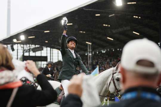 A female rider in a green jacket and white riding breeches sits on a white horse and raises her hand in celebration, waving a white cloth in the air. In the foreground, spectators can be seen waving cloths as well. In the background is a large, covered grandstand filled with people.