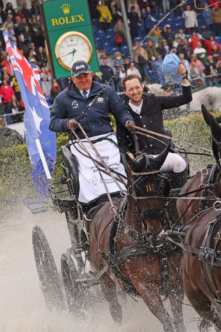 A carriage drawn by several horses drives through a water obstacle. The carriage is decorated with an Australian flag, Martin Fuchs sits next to the driver and holds a blue Rolex cap in the air while water splashes around him. In the background, a large Rolex watch, grandstands and numerous spectators can be seen.