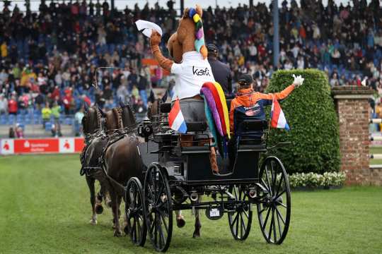 A carriage drawn by four horses drives across the lawn in the main stadium in front of packed stands. Sitting on the carriage is the mascot ‘Karli’ with a colourful mane and tail, cheerfully waving a white cloth. Next to him, a person in orange team clothing waves a Dutch flag.