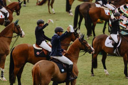 Several riders sit on their horses on the green grass in the main stadium and wave white flags to bid farewell to the nations. More horses and riders can be seen in the background.