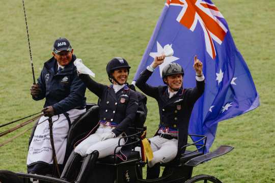 Three people are sitting on a carriage, one of them is driving the carriage and two are wearing tournament clothing and helmets, waving white cloths and cheering. A large Australian flag is flying in the background. The scene takes place on the green lawn in the main stadium.