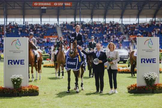 Award ceremony at an equestrian tournament in the stadium: a rider sits on a horse with a blue winner's blanket bearing the RWE logo. Two people are standing next to him, one holding a glass plate, the other a bouquet of flowers. In the background are other riders on their horses and spectators in the stands. Large panels with the CHIO Aachen logo and RWE lettering can be seen on the left and right.