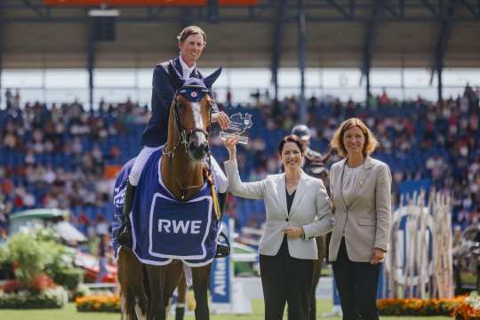 A rider sits on a brown horse wearing a blue winner's blanket with the RWE logo. The rider holds a trophy in his hand and is flanked by two elegantly dressed women on the lawn. In the background is a packed stadium, other horses and large RWE pillars decorated with flowers and the CHIO Aachen logo.