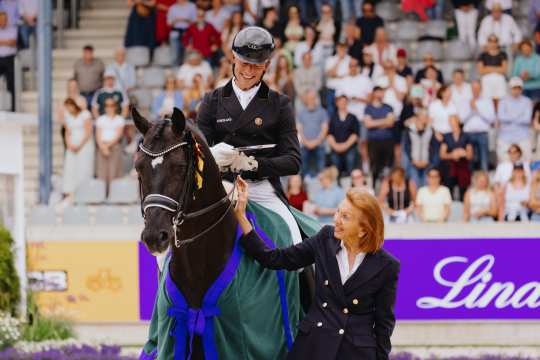 A rider in competition attire sits on a dark brown horse with a blue and green winner's saddle pad. A woman in a dark blazer stands next to the horse and places her hand on the horse's neck. In the background, numerous spectators can be seen in the stands.