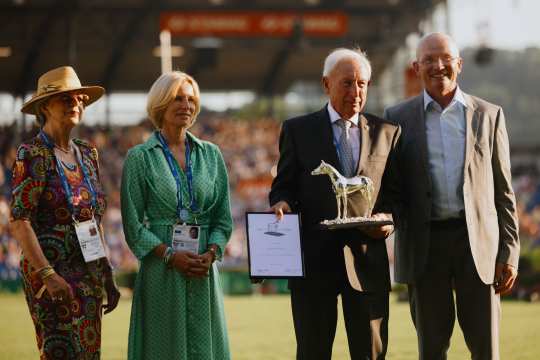 Vier Personen stehen nebeneinander auf einer Wiese im Stadion. Eine Person hält eine silberne Pferde-Statue und eine Urkunde in den Händen. Zwei der Personen tragen bunte Kleider mit Akkreditierungsausweisen, die anderen beiden sind im Anzug gekleidet. Im Hintergrund sind Tribünen mit Publikum zu sehen.