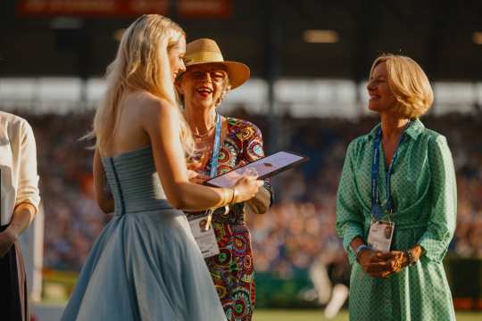 Eine junge Frau im hellblauen Kleid hält lächelnd eine Urkunde in der Hand und steht auf einer Wiese im Stadion. Ihr gegenüber stehen zwei Frauen, eine davon trägt ein buntes Kleid und einen Strohhut, die andere ein grünes Kleid. Im Hintergrund sind unscharfe Tribünen mit Publikum zu sehen.