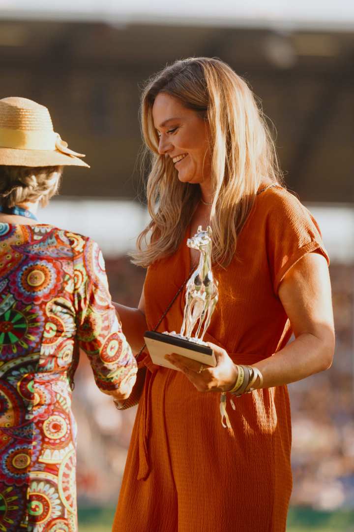 Eine Frau in einem orangefarbenen Jumpsuit hält eine silberne Pferde-Statue als Trophäe in der Hand und steht einer Frau in einem bunten Kleid mit Strohhut gegenüber. Die Szene spielt auf einer Wiese im einem großen Stadion.