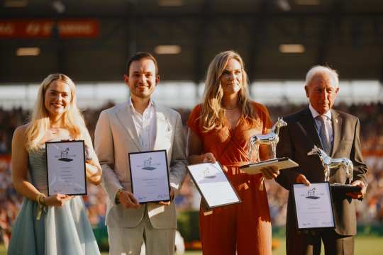 Vier Personen stehen in der Abendsonne nebeneinander in festlicher Kleidung auf einer Wiese im Stadion und halten Auszeichnungen in den Händen. Zwei von ihnen präsentieren zusätzlich silberne Pferde-Statuen als Trophäen. Im Hintergrund sind unscharf gefüllte Tribünen und Stadionbeleuchtung zu sehen.