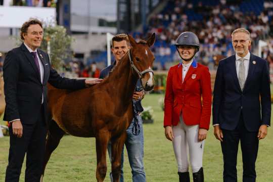 Four people are standing on a meadow in a stadium next to a young brown foal. Two men are wearing suits, one person is holding the foal by the rope, and a female rider in a red jacket and riding helmet is standing next to it. Spectators in the stands can be seen in the background.