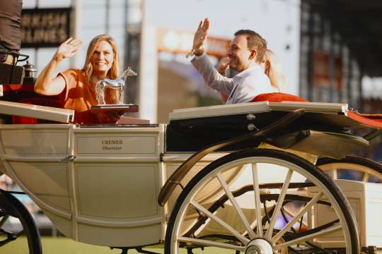 Zwei Personen sitzen in einer hellen, eleganten Kutsche mit der Aufschrift „CREMER Oberzier“. Eine Person hält eine silberne Pferdefigur als Trophäe in der Hand. Beide Personen winken freundlich in die Menge. Die Szene spielt sich auf einer Veranstaltung im Freien ab, im Hintergrund sind unscharfe Tribünen und Schilder zu erkennen.