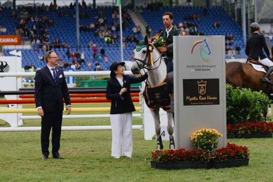 A rider sits on a decorated white horse and holds a bouquet of flowers, while two elegantly dressed people stand next to him. The group is on a jumping arena in front of a large grandstand. In the foreground is a stele with the logo of Mystic Rose Horses and the words “World Equestrian Festival CHIO Aachen”. The horse is wearing a winner's blanket, and further obstacles and another horse can be seen in the background.