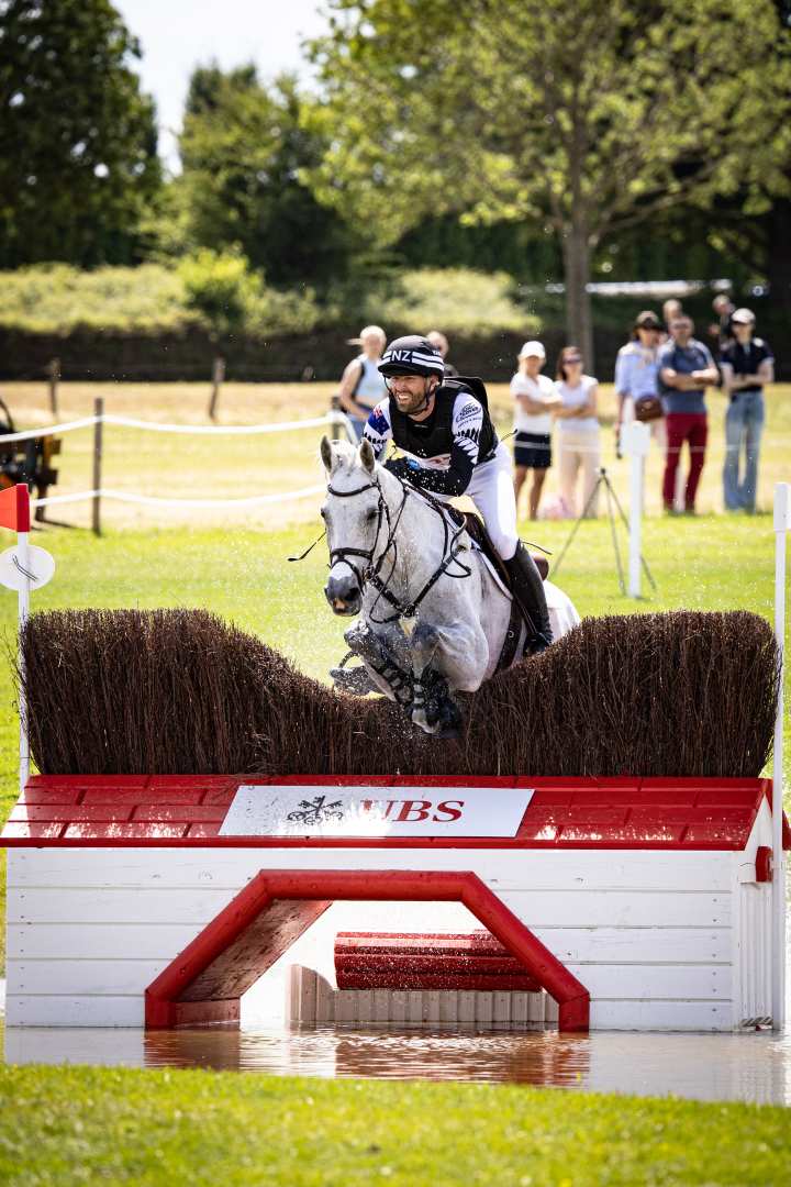 A rider jumps over a water-filled obstacle on a grey horse during an outdoor eventing competition. In the background, several spectators stand on a meadow and watch the competition in sunny weather.