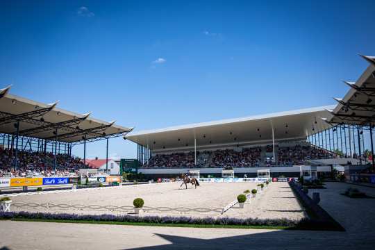 The large Dressage Stadium under a bright blue sky, the stands are filled with numerous spectators. In the foreground is a dressage arena, in the centre of which a rider is riding a brown horse. The dressage stadium is lined with flowers and advertising banners.