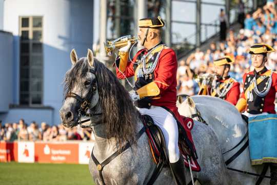 A soldier of the Guardia Civil in a ceremonial uniform with a red jacket, white trousers and gold details sits on a grey horse and blows a golden trumpet. In the background are other horsemen in similar uniforms and numerous spectators in the stands. The scene takes place in sunny weather in the main stadium.