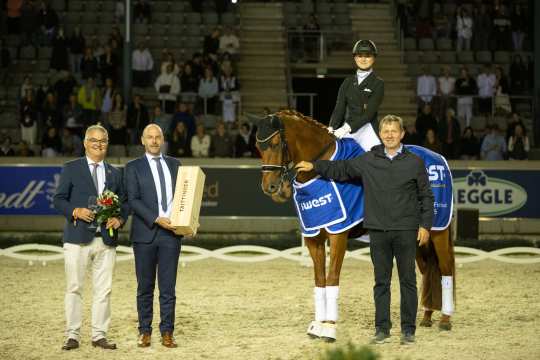 Ein Reiter sitzt auf einem braunen Pferd mit blauer Siegerdecke in einer beleuchteten Reitarena. Neben dem Pferd stehen drei Männer, einer hält einen Blumenstrauß, ein anderer eine große Holzkiste mit der Aufschrift „Taittinger“. Im Hintergrund sind Zuschauer auf den Tribünen zu sehen.
