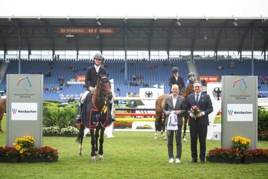A rider sits on a horse adorned with a victory saddle pad and a bow. Next to him are two people, one holding a gift bag, the other a bouquet of flowers. Other riders on horses, obstacles and a large grandstand with rows of blue seats can be seen in the background. To the left and right of the picture are signs with the CHIO Aachen logo and the words “NetAachen”. In front of the signs are floral decorations.