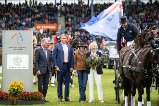 Prize-giving ceremony at the CHIO Aachen: In the foreground, several people are standing on the grass of the main stadium, including an elderly lady with a bouquet of flowers. On the right of the picture is a coachman with a top hat on a team of horses carrying an Australian flag. On the left is a sign with the CHIO Aachen logo and the name "“Familie Richard Talbot”. The full stands of the stadium can be seen in the background.