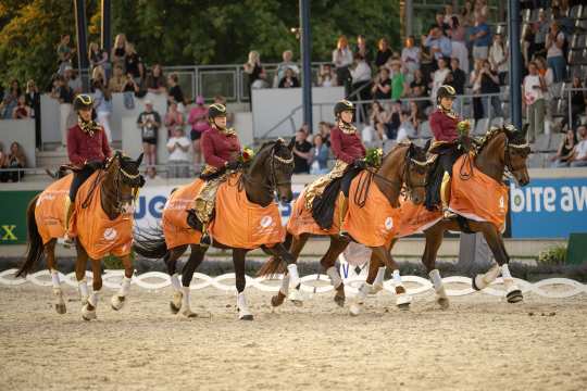 Vier Reiterinnen in festlicher Kleidung reiten nebeneinander auf prächtig geschmückten Pferden, die orangefarbene Siegerdecken tragen. Sie halten Blumensträuße in der Hand und reiten in einer Arena vor gefüllten Tribünen mit applaudierendem Publikum.