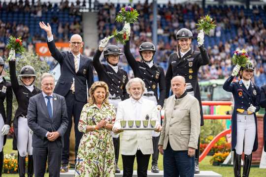 A victorious team in riding gear stands cheering on the podium and holds up bouquets of flowers. In front of them are four people, one of whom is presenting a tray of trophies. Spectators in the stands can be seen in the background.