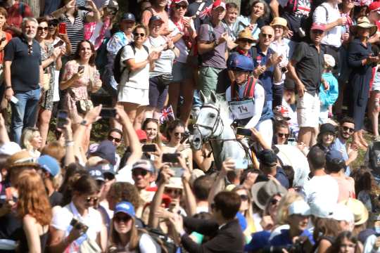 A rider on a white horse rides through a dense crowd at an outdoor event. Many spectators stand and applaud, some hold up cameras or mobile phones, and British flags can be seen in the audience.