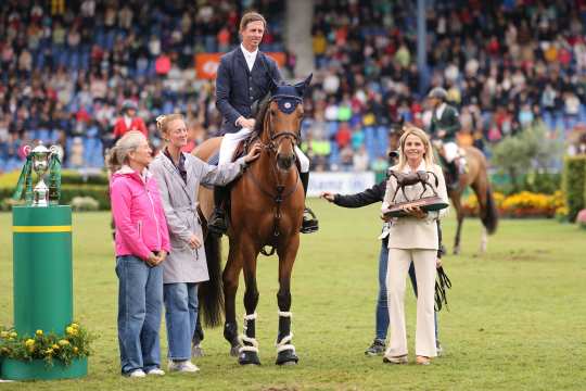 A rider in a dark blue jacket sits on a brown horse and is accompanied by several people on the showground. A woman presents the rider with a horse figurine as a trophy. Other horses, people and spectator stands can be seen in the background.
