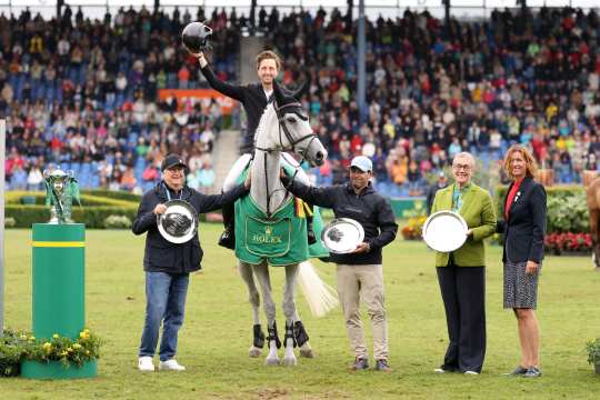 A rider sits on a white horse with a green Rolex victory blanket and raises his helmet in the air in jubilation. Several people with silver plates of honour stand next to him on the lawn, while numerous spectators in the stands applaud in the background.