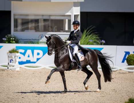 A dressage rider in a black tailcoat and helmet rides a black horse with a white mark on its forehead in the centre of a dressage arena. The horse lifts its legs elegantly in an extended trot; white ribbons with the SAP logo, floral decorations and a judge's house can be seen in the background.