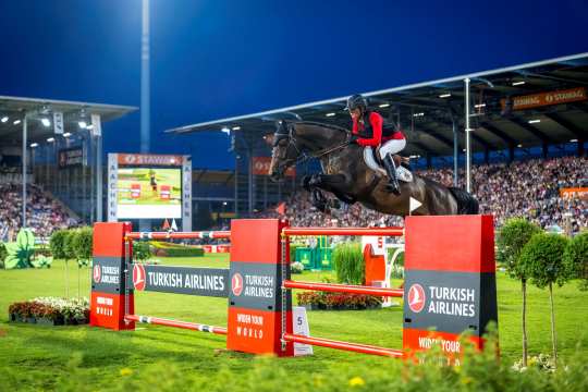 A show jumper in a red jacket and helmet jumps with a dark bay horse over an obstacle with Turkish Airlines branding. The competition takes place in the evening under floodlights, with the stands of a large stadium full of spectators in the background.