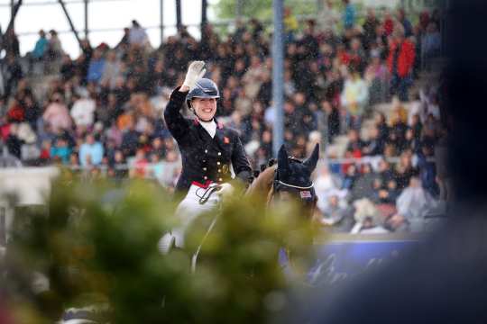 A dressage rider in a dark uniform with gold buttons and a British badge on her chest sits smiling on a brown horse and raises her right hand in greeting. It is raining lightly, in the background many spectators can be seen in the stands, plants in the foreground.
