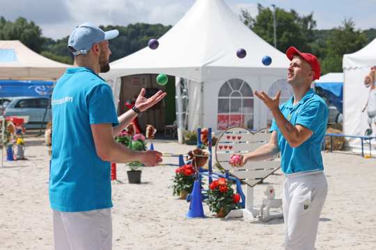 Zwei Männer in blauen T-Shirts und weißen Hosen jonglieren mit bunten Bällen im Sand vor weißen Zelten. Im Hintergrund sind Blumen, Hobby Horsing Hindernisse und ein Herz aus Holz zu sehen.