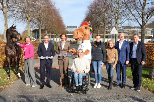 Group photo outdoors in an autumn setting with several people, including a person in a wheelchair, a horse, a person in a horse mascot costume, and others in business and casual attire. Two people are jointly holding a large symbolic key band labeled “Allianz Park.” Leafless trees and a modern building are visible in the background.