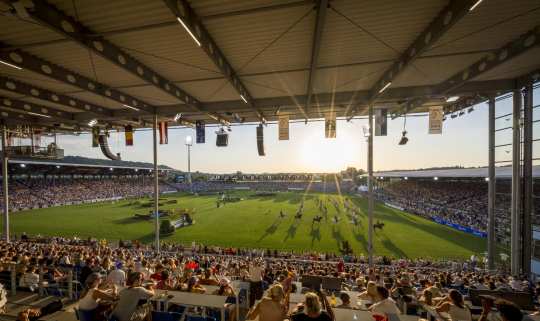 View from a packed covered grandstand in a large stadium at sunset. Horse show performances are taking place on the green lawn, and numerous national flags hang from the grandstand roofs.
