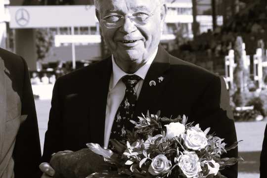 Ein Mann im Anzug und mit gemusterter Krawatte hält einen Blumenstrauß und eine Skulptur in den Händen. Das Bild ist in Schwarz-Weiß gehalten. Im Hintergrund befinden sich eine Reitsportarena und eine große Videowand, die die Preisverleihung zeigt.
