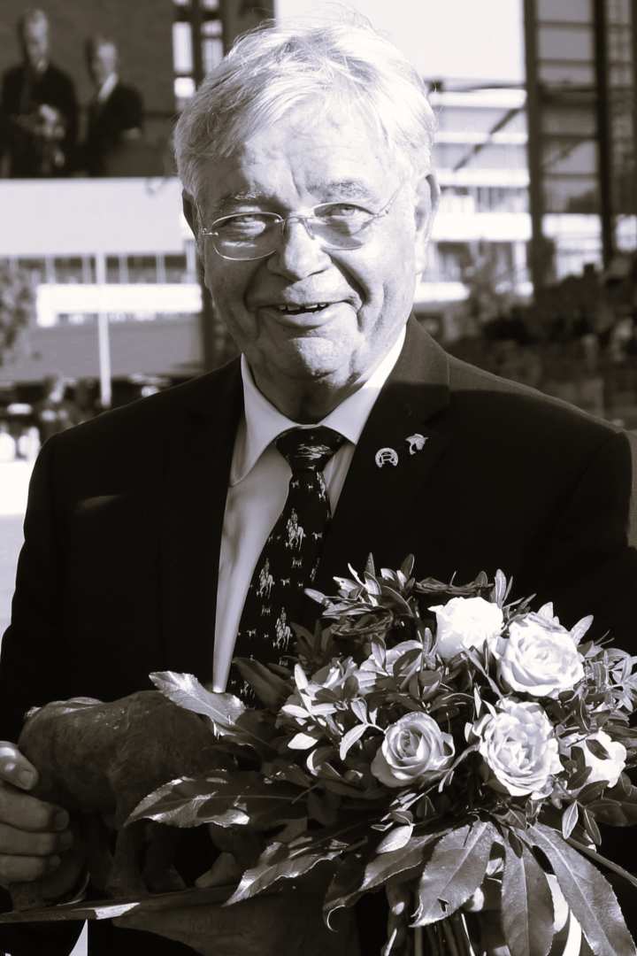 Ein Mann im Anzug und mit gemusterter Krawatte hält einen Blumenstrauß und eine Skulptur in den Händen. Das Bild ist in Schwarz-Weiß gehalten. Im Hintergrund befinden sich eine Reitsportarena und eine große Videowand, die die Preisverleihung zeigt.