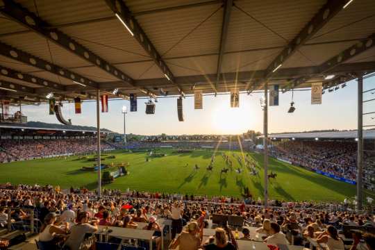 Blick aus einer vollbesetzten überdachten Tribüne in ein großes Stadion bei Sonnenuntergang. Auf dem grünen Rasen finden Showvorführungen mit Pferden statt, zahlreiche Nationenflaggen hängen von den Tribünendächern.