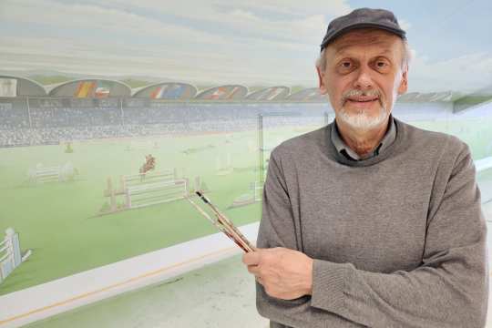 Ein älterer Mann mit grauem Pullover und schwarzer Kappe steht vor einer Wandmalerei mit einer Stadion- und Springreiterszene und hält mehrere Pinsel in der Hand.