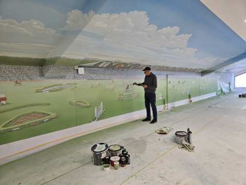 In einem langen Innenraum bemalt ein Künstler eine großflächige Wandmalerei mit einer Stadion- und Reitsportszene unter blauem Himmel. Auf dem Boden stehen Farbeimer und Pinsel, rechts fällt Licht durch ein Fenster in den Raum.