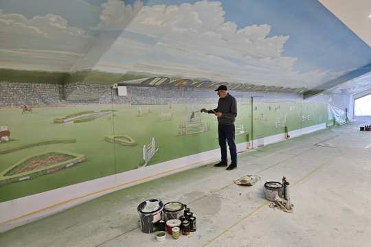 In einem langen Innenraum bemalt ein Künstler eine großflächige Wandmalerei mit einer Stadion- und Reitsportszene unter blauem Himmel. Auf dem Boden stehen Farbeimer und Pinsel, rechts fällt Licht durch ein Fenster in den Raum.