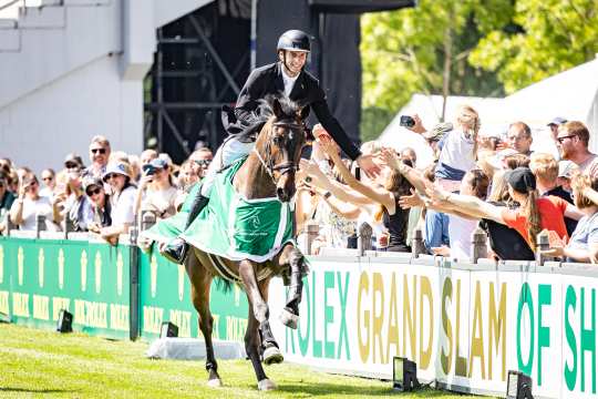 Rider in a black jacket and helmet celebrates on a brown horse wearing a green winner’s blanket, reaching out to high-five spectators along the arena rail; behind them a cheering crowd and boards with the lettering “ROLEX GRAND SLAM OF SHOW JUMPING” on a sunny day.