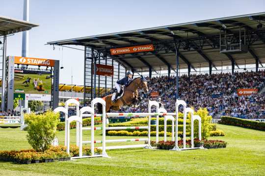 Springreiterin auf einem fuchsfarbenen Pferd springt über ein hohes, weißes Hindernis im vollbesetzten Stadion beim CHIO Aachen, im Hintergrund Videowand und Werbebanner von STAWAG und Rolex.