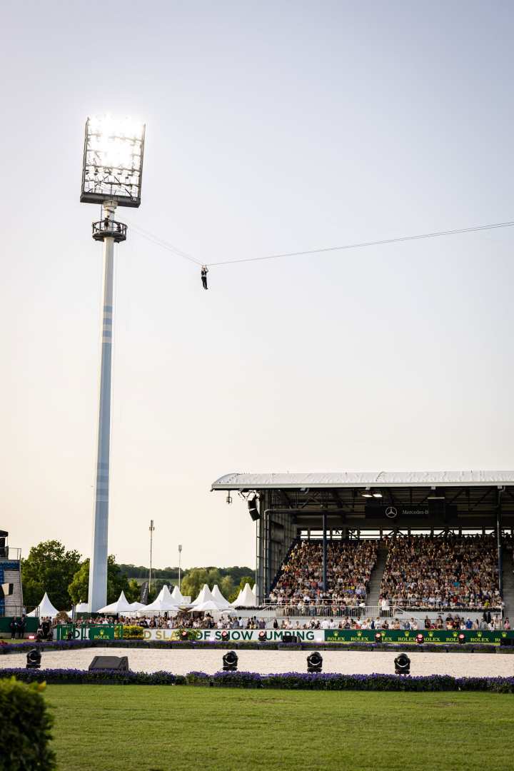 Weitwinkelaufnahme im Reitstadion: Hoch oben an einer Zipline hängt eine Person an einem Seil vor hellem Himmel, darunter ist eine voll besetzte Tribüne und die Arena mit „Rolex“-Banden zu sehen.