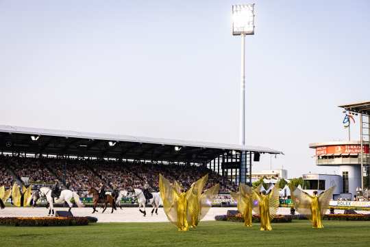 An evening shot of the stadium in Aachen: Dancers in golden winged costumes stand on the field, while riders on horseback pass by in the background; the stands are packed, and a “Turkish Airlines” tower can be seen on the right.