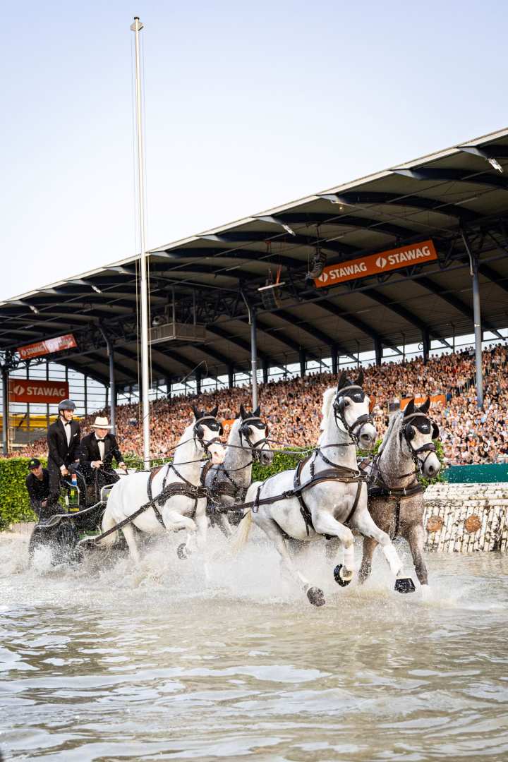 Ein Vierspänner mit vier weißen Pferden spritzt durch ein Wasserhindernis im Stadion; die Fahrer tragen Frack und Helm, im Hintergrund ist die voll besetzte Tribüne mit STAWAG-Bannern zu sehen.