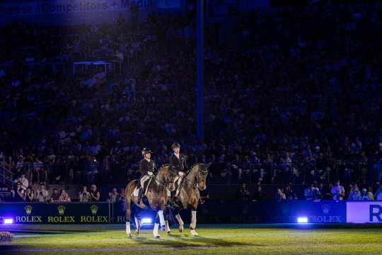 Two riders are riding through the stadium under the floodlights; in the background, a large crowd sits bathed in blue light, and “Rolex” logos are visible several times along the boards.
