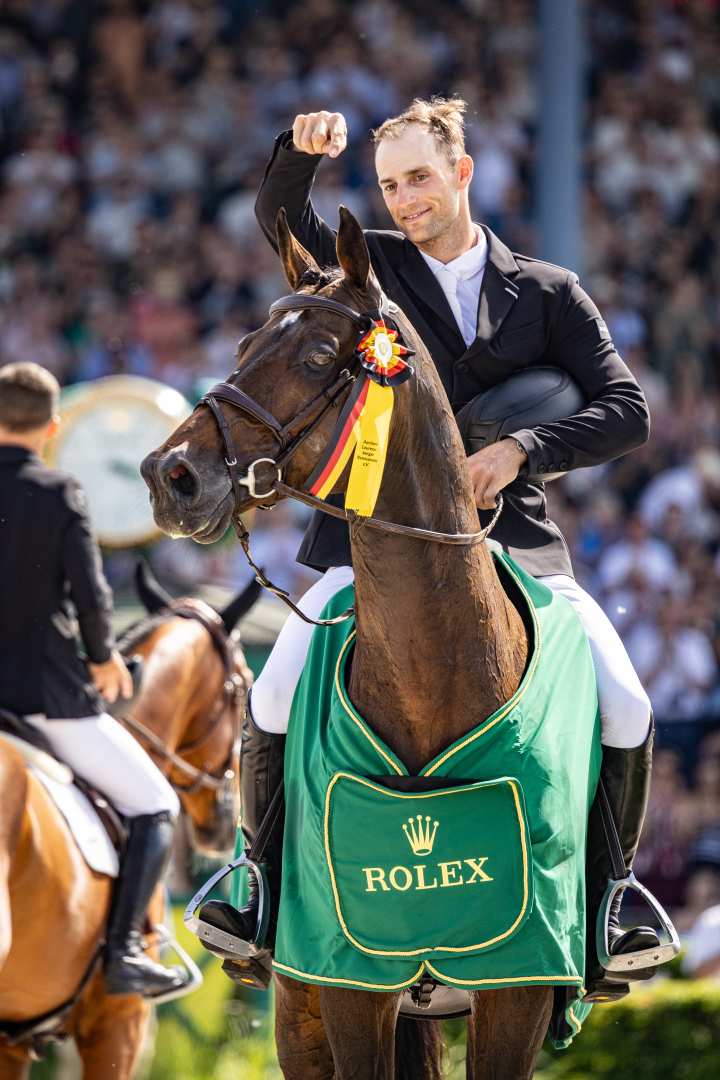 A show jumper in a black blazer sits on a bay horse draped in a green Rolex blanket and raises his fist in celebration; a black, red, and yellow ribbon hangs from the bridle, and the crowd can be seen in the background, blurred.
