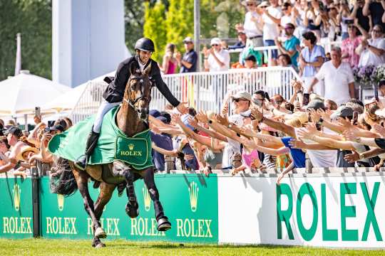 A show jumper in a black blazer rides a bay horse draped in a green Rolex blanket along the track, reaching out his hand to cheering spectators who are clapping and taking photos of him.