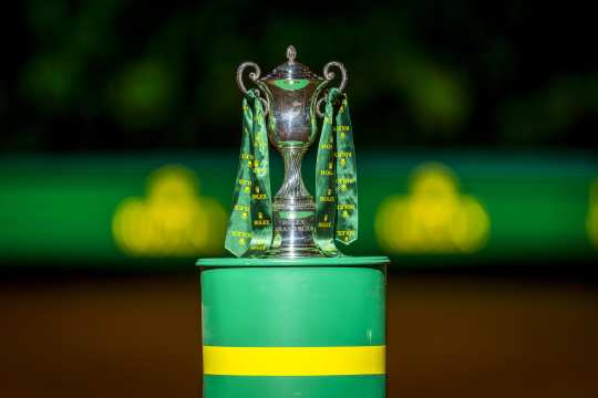 Ein silberner „Rolex Grand Slam“-Pokal mit grünen Bändern steht auf einem grün-gelben Podest vor unscharfem, dunkelgrünem Hintergrund.