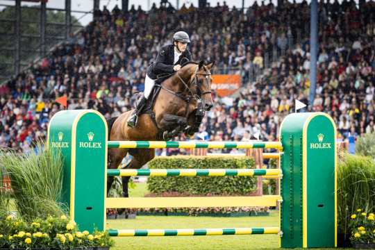 Springreiter auf einem braunen Pferd springt im Regen über ein grün-gelbes Rolex-Hindernis in einem Stadion mit voll besetzten Zuschauerrängen.