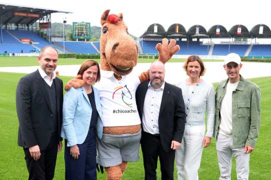 Gruppe von vier Frauen und Männern steht auf dem Rasen eines großen Reitstadions neben einem Pferdemaskottchen in braunem Fellkostüm. Das Maskottchen trägt ein weißes T Shirt mit buntem Pferdelogo und der Aufschrift „#chioaachen“ und breitet einen Arm winkend aus. Im Hintergrund sind leere blaue Tribünen, ein Springplatz und mehrere Nationalflaggen zu sehen.