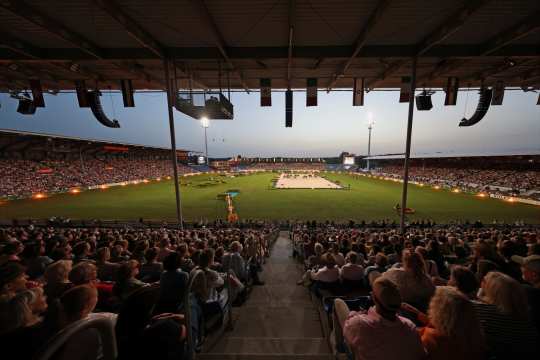 Wide-angle view of an evening equestrian stadium with packed grandstands in the foreground; in the brightly lit grass arena a sand-colored dressage ring with several horses and riders is visible, surrounded by floodlights and national flags hanging from the roof.